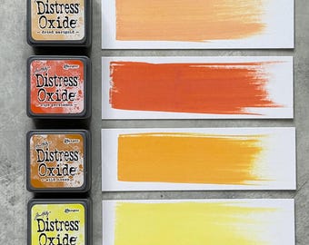 Può includere: Quattro tamponi di inchiostro Distress Oxide in calendula essiccata, kaki maturo, miele selvatico e limonata spremuta. Ogni tampone è accanto a una corrispondente campionatura di colore su carta bianca. I tamponi di inchiostro sono etichettati con il marchio e il colore.