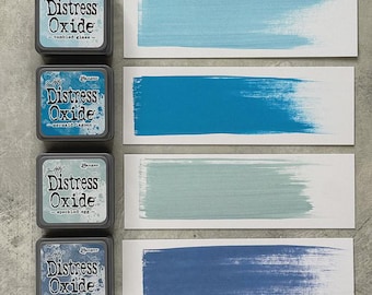 Può includere: Quattro tamponi di inchiostro Distress Oxide in tonalità blu e verde, con campioni di colore corrispondenti. I tamponi di inchiostro sono etichettati "tumbled glass", "mermaid lagoon", "speckled egg" e "faded jeans". Ogni campione mostra una pennellata del colore.