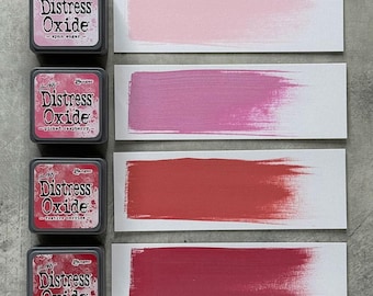 Può includere: Quattro tamponi di inchiostro Distress Oxide in tonalità rosa e rosse, con campioni di colore corrispondenti. I tamponi di inchiostro sono etichettati "spun sugar", "picked raspberry", "festive berries" e "lumberjack plaid". Ogni campione mostra una pennellata del colore dell'inchiostro.