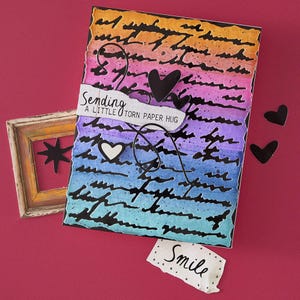 May include: A handmade greeting card with a gradient background of orange, pink, purple, and blue. The card features black script, black heart cutouts, and the text "Sending a little torn paper hug." A small piece of paper says "Smile."