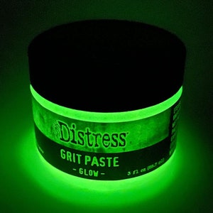 May include: A round black container of Distress Grit Paste - Glow. The container has a black lid and a glowing green band around the middle. The label reads "Distress Grit Paste - Glow" and the container holds 3 fl oz (88.7 ml).