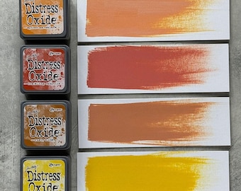 Puede incluir: Cuatro almohadillas de tinta Distress Oxide en tonos naranja, rojo y amarillo, con muestras de color correspondientes. Las almohadillas de tinta están etiquetadas como "spiced marmalade", "crackling campfire", "rusty hinge" y "mustard seed". La tinta se aplica sobre papel blanco.