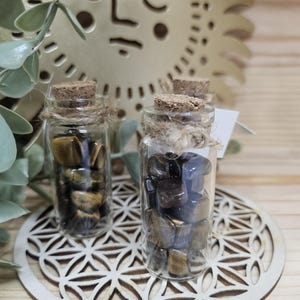 Puede incluir: Pequeñas botellas de vidrio llenas de piedras preciosas de ojo de tigre, cada una sellada con un corcho y cuerda. Las botellas están sobre una superficie de madera con un diseño geométrico decorativo. Un recorte dorado en forma de sol está en el fondo.