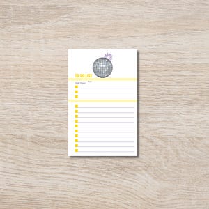 以下が含まれることがあります： 「TO DO LIST」と書かれた黄色のヘッダーが付いた白いメモ帳。 メモ帳には、ディスコボールのグラフィックとメモ用の罫線スペースがあります。 メモ帳は長方形で、明るい木製の表面にあります。