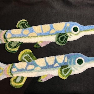 Peut inclure: Deux tapis faits à la main en forme de poisson. Chaque tapis présente un corps long et élancé avec un motif bleu et crème, des nageoires vertes et un grand œil vert. Les poissons sont posés sur un fond sombre.