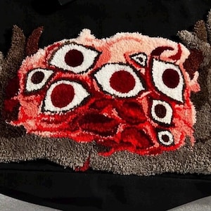 May include: A black tote bag featuring a tufted, textured patch with a central cluster of multiple white and dark red eyes surrounded by shades of pink, red, and brown yarn. The design includes organic, flowing shapes in grey and brown fibers.