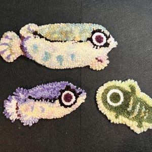 May include: Three handmade, textured fish-shaped decorative items. The largest is off-white with blue and purple accents, the middle one is purple and yellow, and the smallest is green and yellow. Each has a dark eye.