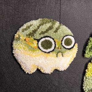 May include: A circular, textured rug with a cartoon face design. The rug is in shades of white, yellow, and green, with black and white detailing for the eyes and glasses. The rug has a whimsical, playful aesthetic, suitable for a child's room.