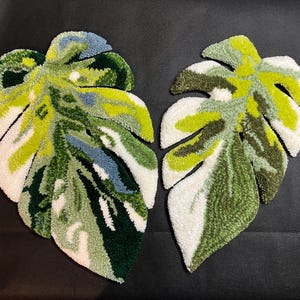 May include: Two leaf-shaped decorative rugs. Each rug features a monstera leaf design with a mix of green, white, and blue hues. The rugs are made of a textured material, likely yarn or fabric, and are placed on a dark surface.