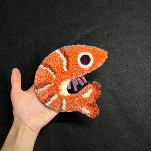May include: A punch needle textile art piece shaped like a shrimp, featuring vibrant orange and white yarn textures, a dark purple circular eye, and small purple yarn details representing the legs on a curved body.