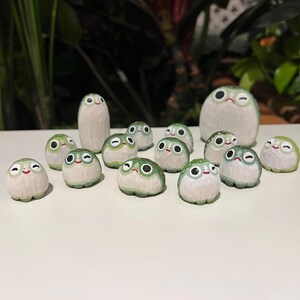 May include: A collection of hand-carved wooden frog figurines. The frogs have white bodies with green accents and expressive faces. Some are round, others are taller. The collection is arranged on a white surface with greenery in the background.