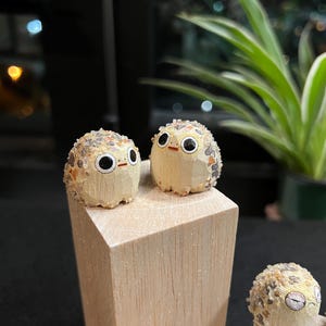 Puede incluir: Dos pequeñas figuras redondas de madera con grandes ojos negros y pequeños picos naranjas. Las figuras tienen un patrón texturizado y moteado en la cabeza y la espalda. Se exhiben sobre un pequeño bloque de madera clara.