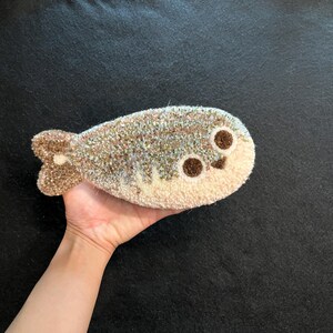 May include: A punch needle textile art piece shaped like a fish, featuring a textured surface with a blend of beige, brown, and light blue yarn. The fish has two dark brown circular eyes and a distinct tail fin, crafted with a dense loop pile.