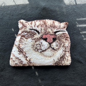 May include: A tufted rug shaped like a cat head with a smiling expression. The rug features a textured surface with a color palette of white, cream, and various shades of brown, including a distinct pink nose and closed eyes in a curved shape.