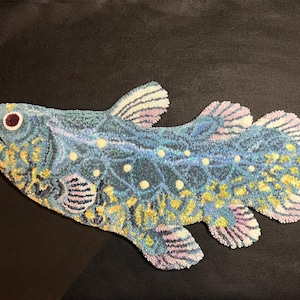 May include: A tufted textile wall hanging shaped like a coelacanth fish. The surface features a textured loop pile in shades of blue, yellow, and white, with a dark red eye detail and light purple accents on the fins and tail edges.