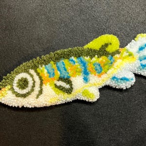 Puede incluir: Una colorida alfombra hecha a mano con forma de pez. La alfombra presenta una base blanca con detalles en verde, amarillo, azul y naranja. El pez tiene un ojo y aletas detallados, y est&aacute; colocado sobre una superficie oscura. Esta alfombra decorativa es un art&iacute;culo de decoraci&oacute;n &uacute;nico.