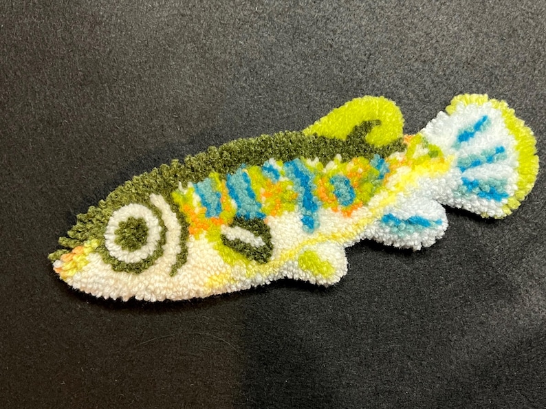 Puede incluir: Una colorida alfombra hecha a mano con forma de pez. La alfombra presenta una base blanca con detalles en verde, amarillo, azul y naranja. El pez tiene un ojo y aletas detallados, y est&aacute; colocado sobre una superficie oscura. Esta alfombra decorativa es un art&iacute;culo de decoraci&oacute;n &uacute;nico.