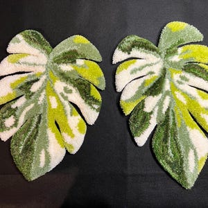 May include: Two leaf-shaped rugs with a monstera leaf design. The rugs feature a tufted texture and a color palette of white, green, and yellow. The leaves are arranged on a black background.