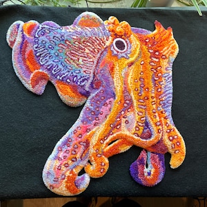 Op de afbeelding: Een getuft tapijt in de vorm van een zeekat, met een levendig kleurenpalet van oranje, paars, roze en wit. Het ontwerp toont ingewikkelde, getextureerde patronen over de mantel en tentakels, met een opvallend donker cirkelvormig oog.