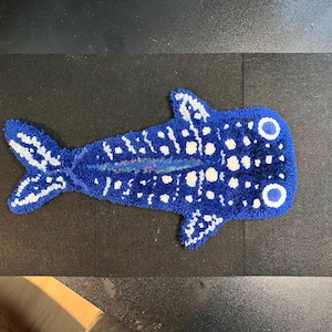 May include: A blue whale shark rug with white spots and accents. The rug is shaped like a whale shark, with a tail and fins. The rug is made of a soft, textured material.