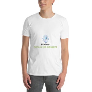 Può includere: T-shirt bianco con grafica di un volto di robot stilizzato in blu e verde, con la scritta "AI is here. humans still debugging" in verde e nero. La maglietta è a maniche corte e sembra essere di cotone.