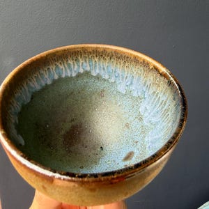 May include: A handmade ceramic bowl with a unique glaze. The bowl features a brown exterior, transitioning to a light blue and green interior. The rim has a textured, dripped glaze effect. This bowl is ideal for serving food or as a decorative piece.
