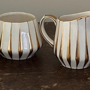 May include: A white and gold striped creamer and sugar bowl set. The creamer has a curved handle and a pouring spout. The sugar bowl is round with a wide opening. Both pieces have gold accents.