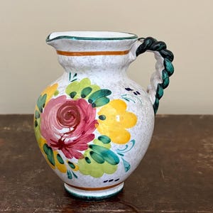 Peut inclure: Un petit pichet en céramique blanche avec une anse tressée. Le pichet est décoré de motifs floraux colorés en rose, jaune et vert. Le bord et la base sont ornés de bandes vertes et blanches. La poignée est de couleur vert foncé.