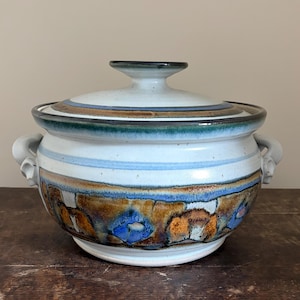 May include: A ceramic lidded casserole dish featuring a light gray base with horizontal bands of blue and brown glaze. The lower section displays a textured pattern of brown and blue shapes. It includes two side handles and a matching lid.