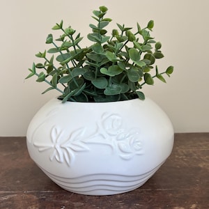 May include: A white ceramic vase with embossed floral patterns, containing a vibrant green plant. The vase is round with a textured surface and is filled with a lush, leafy plant, creating a decorative accent.
