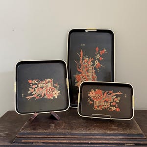 May include: A set of three black serving trays with floral and cart designs. Each tray has a gold trim and woven handles. The designs feature red, gold, and white flowers and cart motifs, creating a vintage aesthetic.