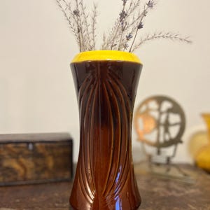 May include: A brown ceramic vase with a yellow rim, containing dried lavender. The vase features a textured, vertical pattern. The vase is on a wooden surface.