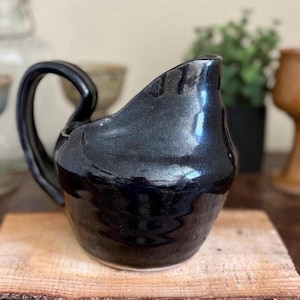 May include: A black ceramic jug with a curved handle. The jug has a glossy finish and sits on a wooden surface. The design is simple and elegant, suitable for serving beverages. The jug is likely handmade.