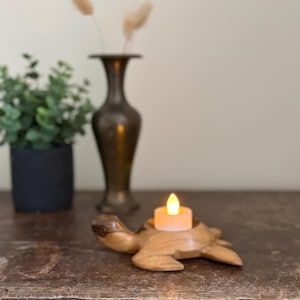 May include: A wooden turtle-shaped candle holder with a lit, battery-operated candle. The candle holder is light brown and sits on a dark wooden surface. A black pot with greenery and a bronze vase are in the background.