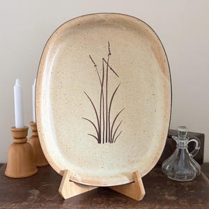May include: Oval ceramic serving platter with a speckled beige surface and a dark brown plant design. The platter is displayed on a wooden stand. A glass cruet and candles in wooden holders are also visible.