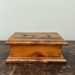 May include: A small, rectangular wooden box with a hinged lid. The box is made of light brown wood and features a dark silhouette of a horse-drawn carriage on the lid. The box has a simple, rustic design.