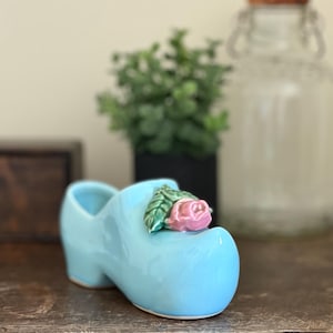 May include: A light blue ceramic clog with a pink rose and green leaves on the front. The decorative shoe is a miniature version of a traditional Dutch wooden shoe. The clog is displayed on a wooden surface.
