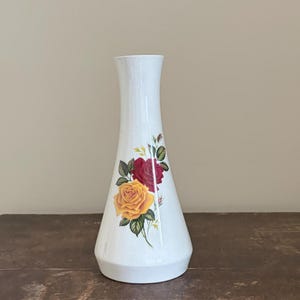 May include: White ceramic vase with a flared base and a slender neck. The vase is decorated with a floral design featuring a yellow and red rose with green leaves. The vase is approximately 9 inches tall.