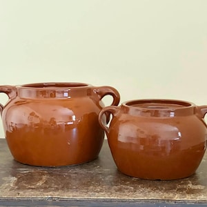 Peut inclure: Deux pots en céramique bruns avec des corps arrondis et des anses en boucle. Les pots ont une finition brillante et reposent sur une surface en bois. Le plus grand pot est à gauche et le plus petit à droite.