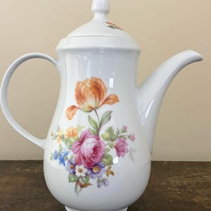 May include: White porcelain coffee pot with a lid and handle. The pot is decorated with a floral design featuring an orange tulip, pink roses, and other colourful flowers. The spout is curved, and the pot sits on a round base.