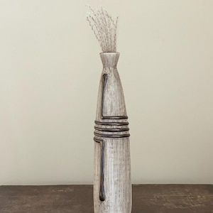 May include: A tall, slender ceramic vase with a textured, off-white surface featuring vertical etched lines. The vase is accented with dark brown, smooth horizontal bands and vertical detailing. A small bunch of dried, light-coloured grass is inside.