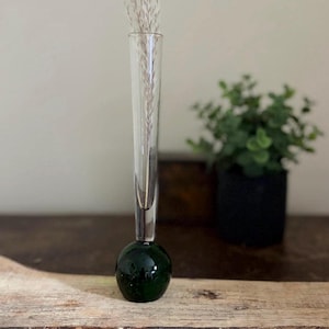 May include: A clear glass bud vase with a dark green, spherical base. The vase holds dried ornamental grass. The vase is on a wooden surface, with a small potted plant in the background.