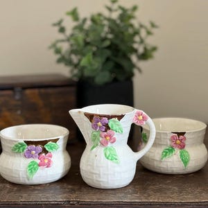 Peut inclure: Un ensemble de pot à crème et sucrier en céramique blanche avec un motif de tissage en panier. L'ensemble est orné de fleurs roses et violettes et de feuilles vertes. Le pot à crème a une anse et un bec verseur.