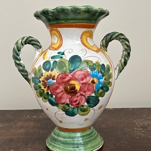 May include: A hand-painted ceramic vase with a white base, decorated with colourful floral designs. The vase has a green rim and base, with twisted green handles. The flowers are red, blue, and yellow, with green leaves. A decorative item for the home.