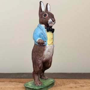 May include: A ceramic rabbit figurine, standing upright on a green base. The rabbit is brown with white ears and wears a light blue jacket, yellow vest, and black bow tie. A decorative Easter or spring-themed item.