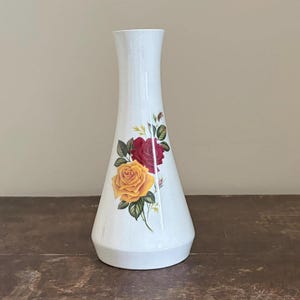 May include: White ceramic vase with a flared base and a narrow neck. The vase is decorated with a floral design featuring a yellow and a red rose with green leaves. The vase is on a wooden surface.