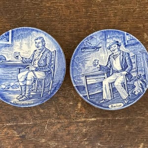 May include: Two blue and white ceramic coasters with illustrations of men. One coaster depicts a man labeled "Huntsman" seated at a table, and the other shows a man labeled "Sailor" in a similar setting. Both are holding a glass.