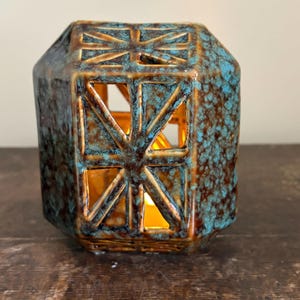May include: A ceramic teal and brown geometric lantern with cut-out starburst designs. The lantern has a textured surface and a warm glow emanating from within, suggesting a candle is lit inside.