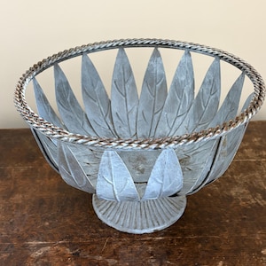 May include: A decorative metal bowl with a leaf design. The bowl is painted in a muted gray color and features a twisted metal rim. The bowl sits on a round base and is suitable for displaying fruit or other items.
