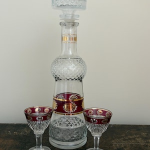May include: A clear glass decanter with a stopper, and two matching cordial glasses. The decanter has a red and gold band around the middle. The cordial glasses have a red rim with gold accents. The set is on a wooden surface.